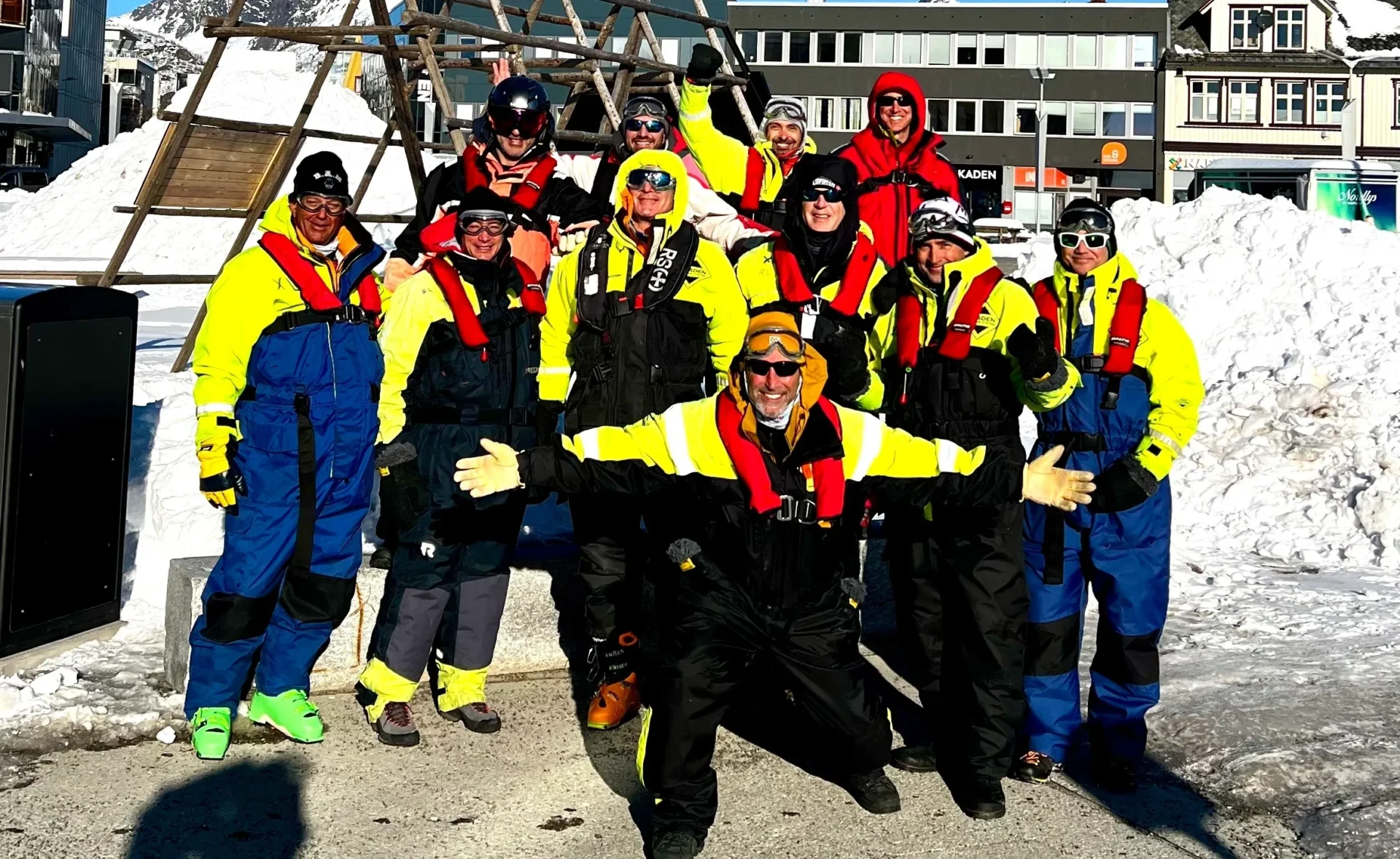 Group of skiers posing on a snowy mountain with lake and peaks behind.