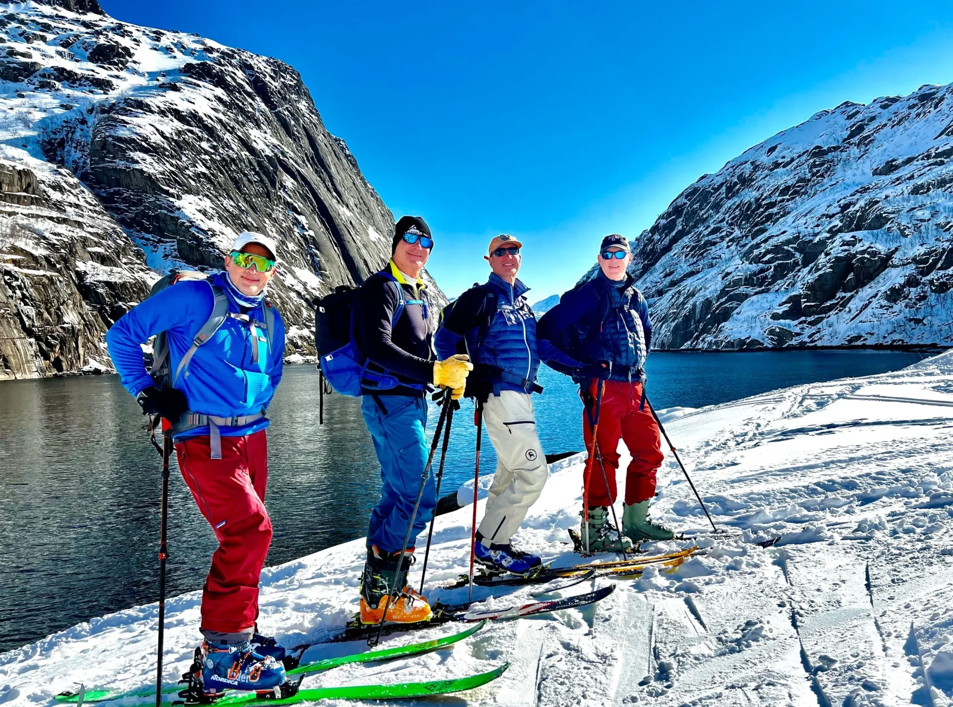 Group of skiers posing on a snowy mountain with lake and peaks behind.