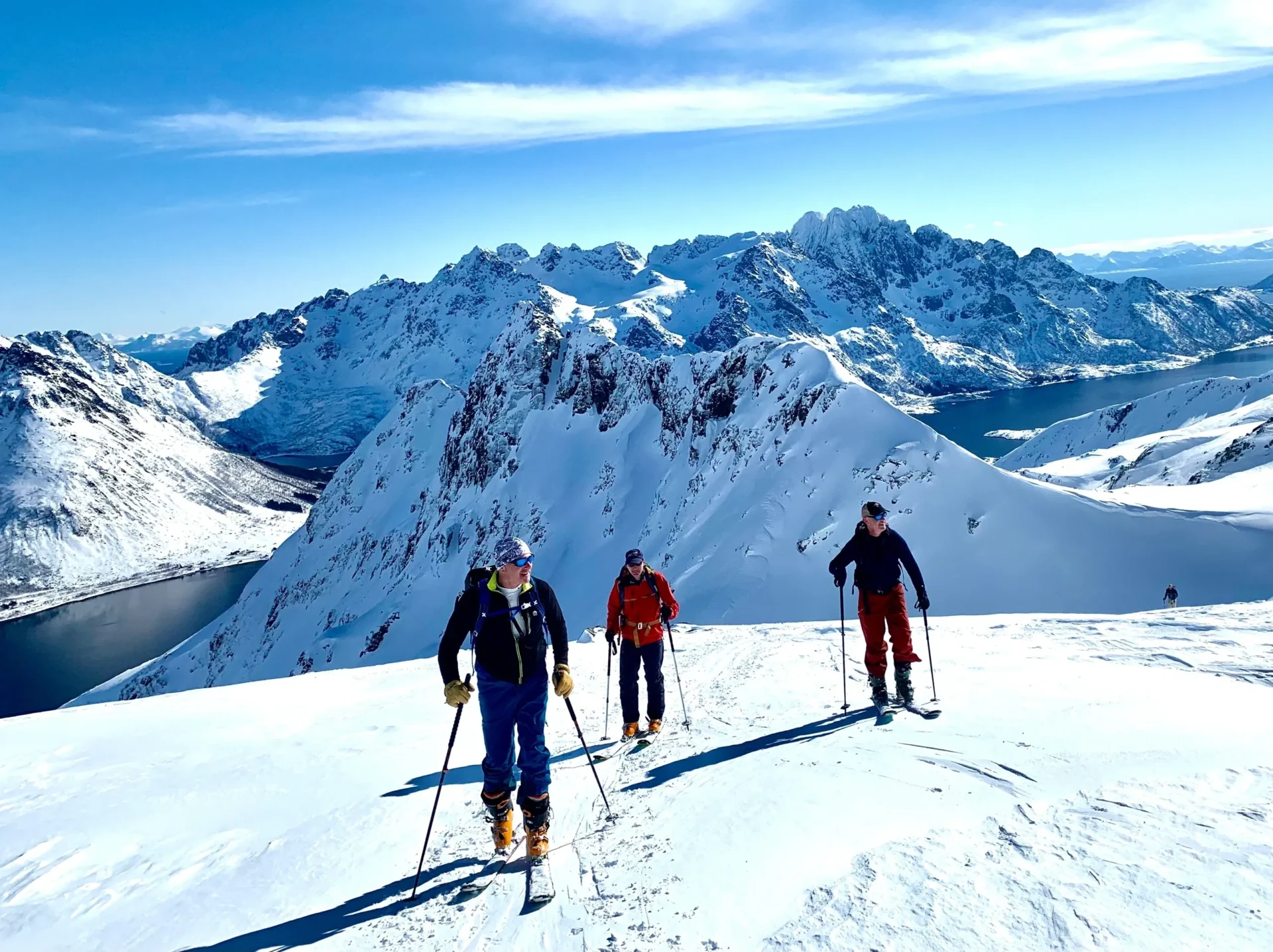Group of skiers posing on a snowy mountain with lake and peaks behind.