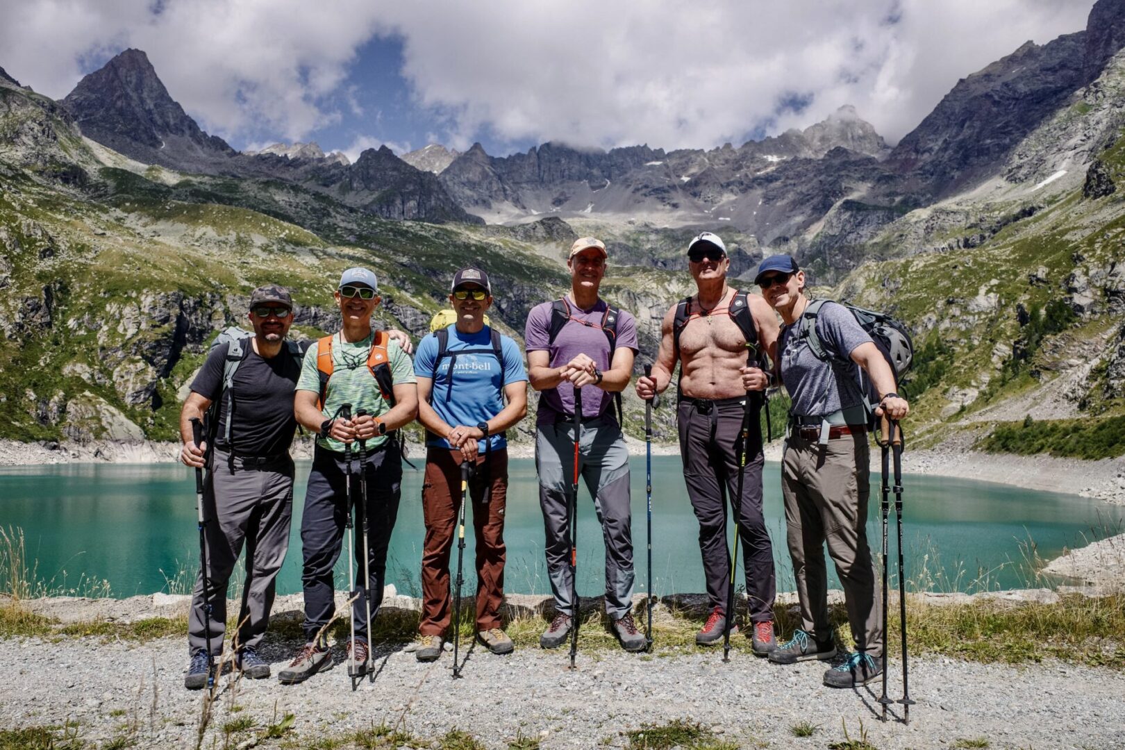 Group of skiers posing on a snowy mountain with lake and peaks behind.
