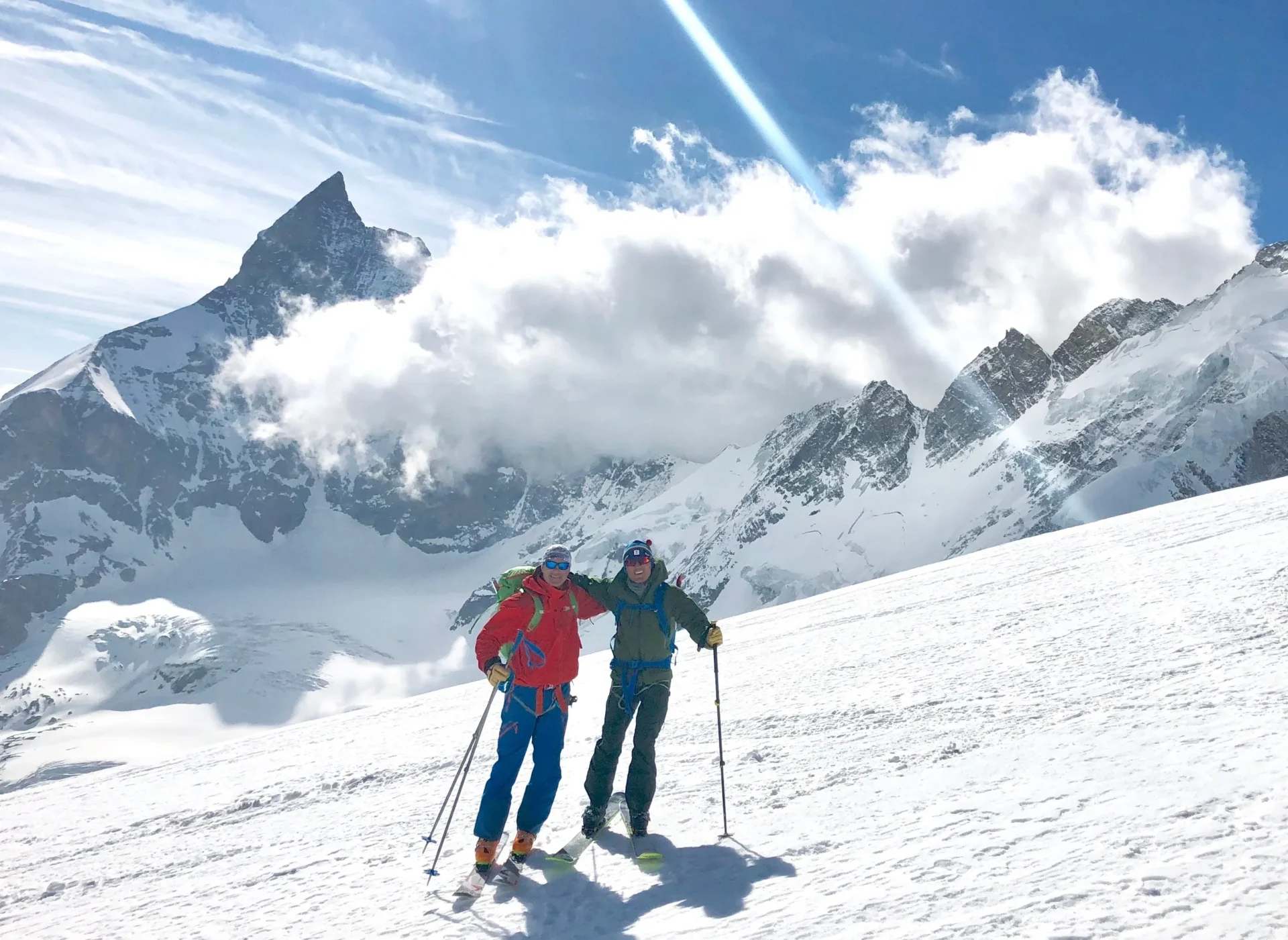 Group of skiers posing on a snowy mountain with lake and peaks behind.