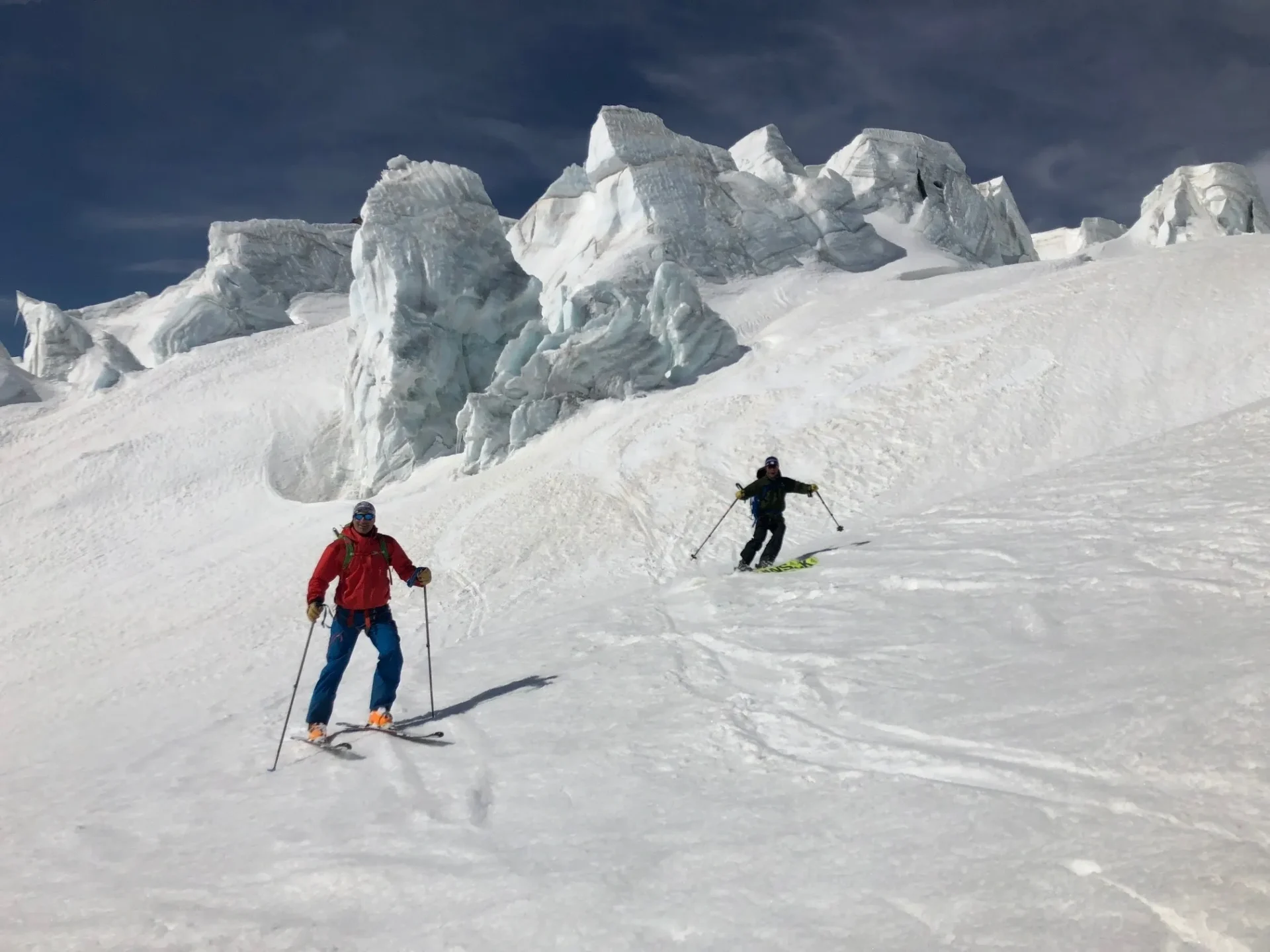 Two skiers descend a snowy mountain with icy peaks in the background.