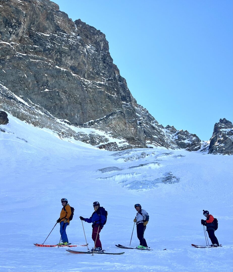 Group of skiers posing on a snowy mountain with lake and peaks behind.