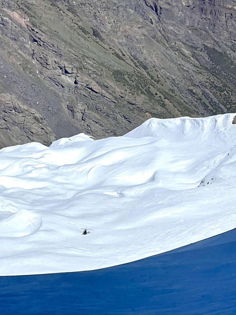 Group of skiers posing on a snowy mountain with lake and peaks behind.