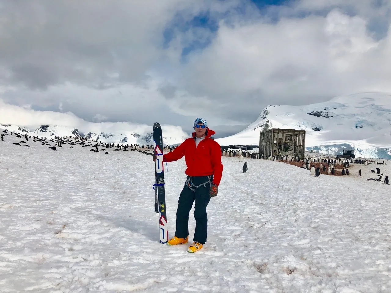 Group of skiers posing on a snowy mountain with lake and peaks behind.