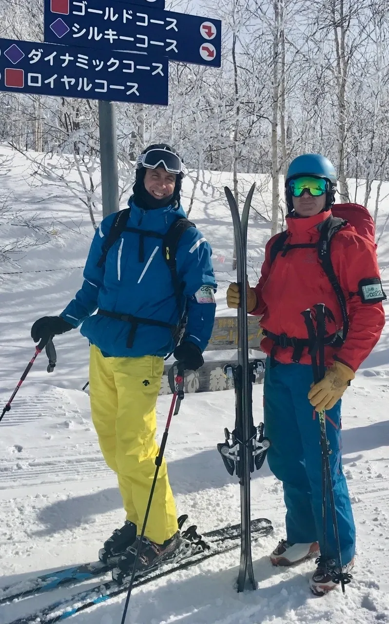 Group of skiers posing on a snowy mountain with lake and peaks behind.