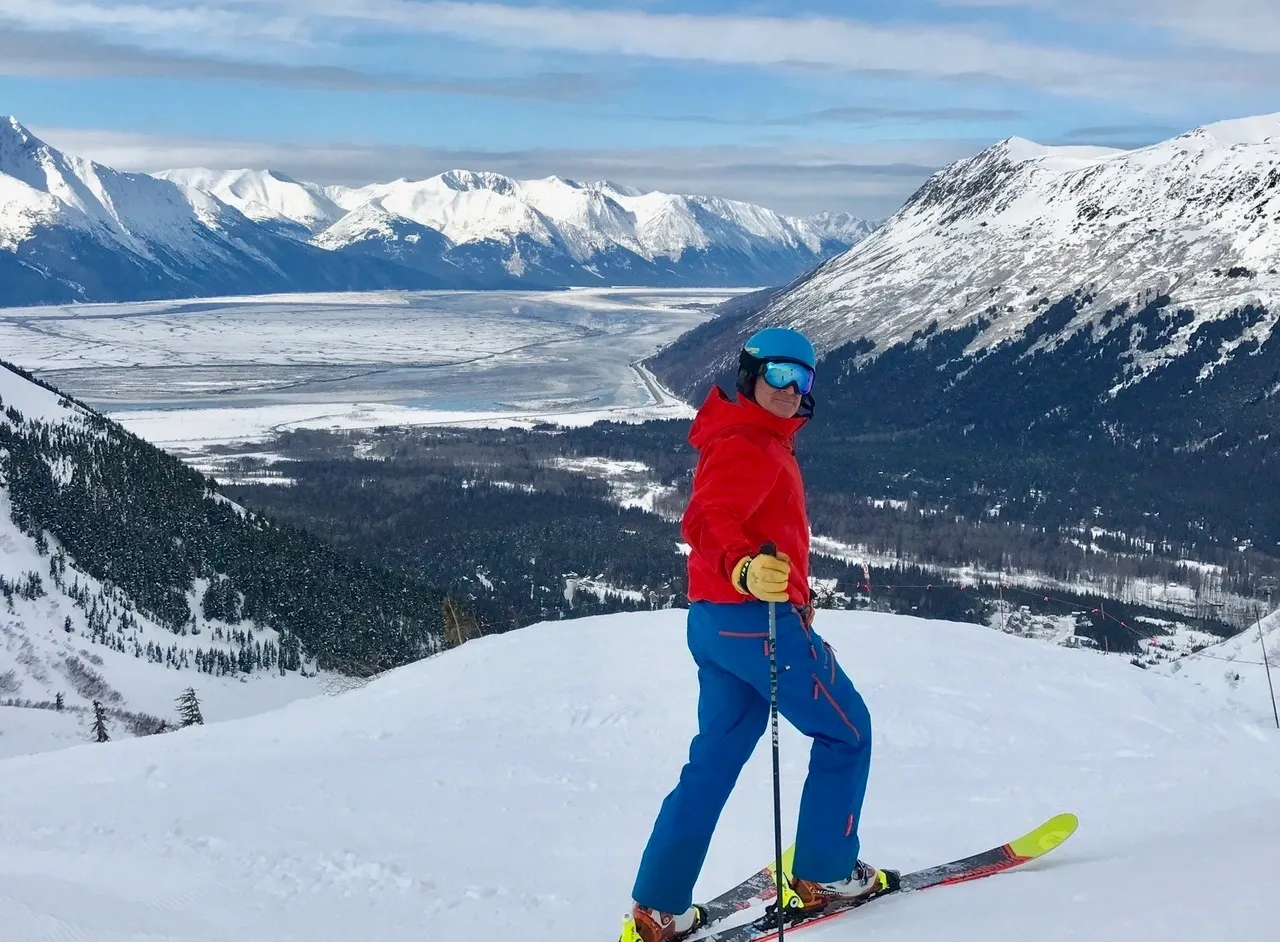 Group of skiers posing on a snowy mountain with lake and peaks behind.