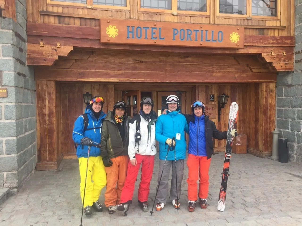 Group of skiers posing on a snowy mountain with lake and peaks behind.