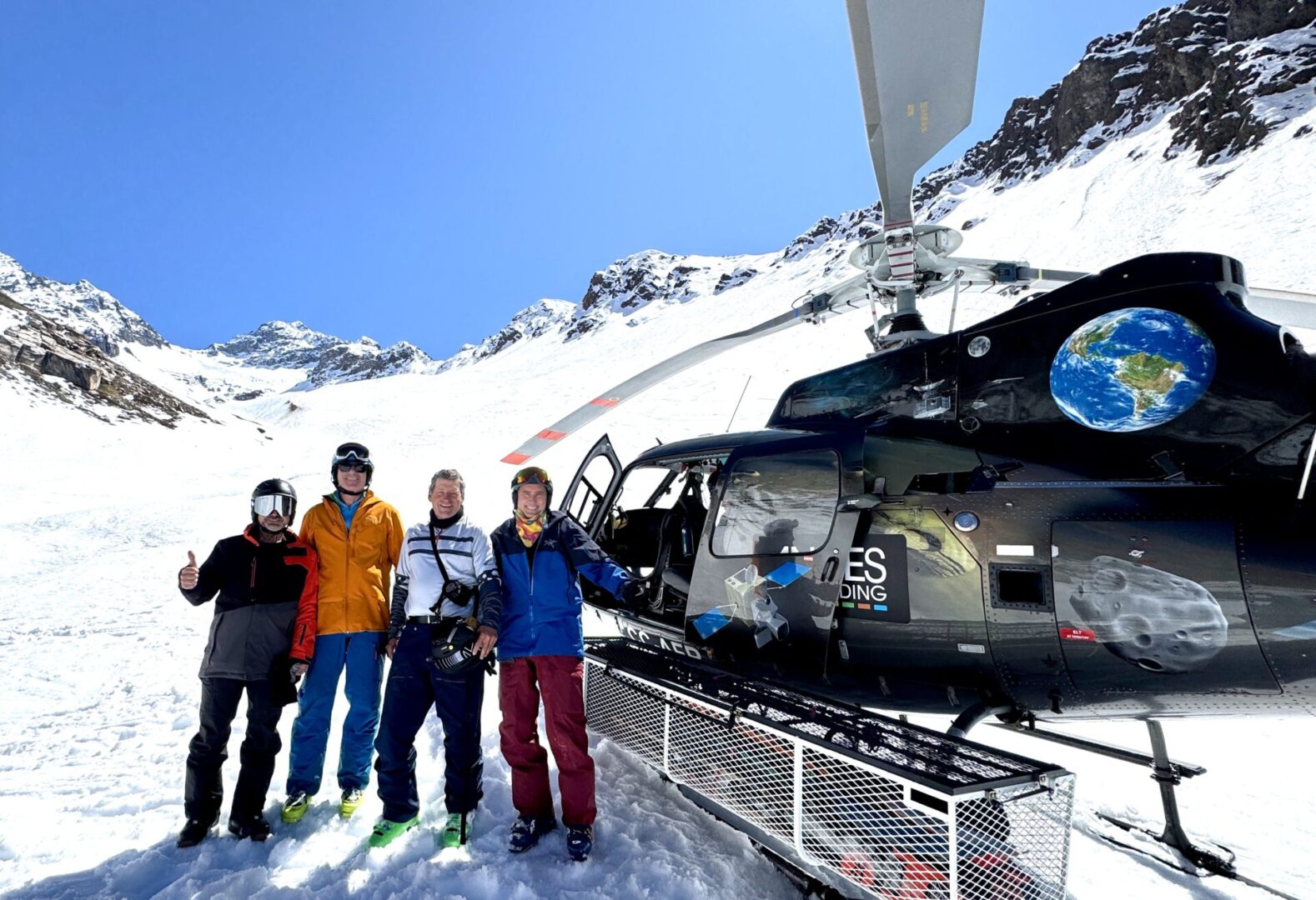 Group of skiers posing on a snowy mountain with lake and peaks behind.