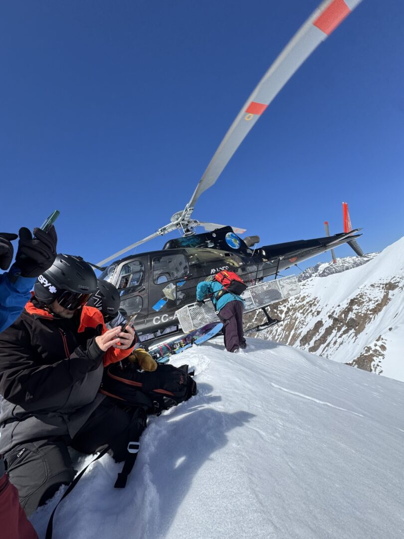 Group of skiers posing on a snowy mountain with lake and peaks behind.
