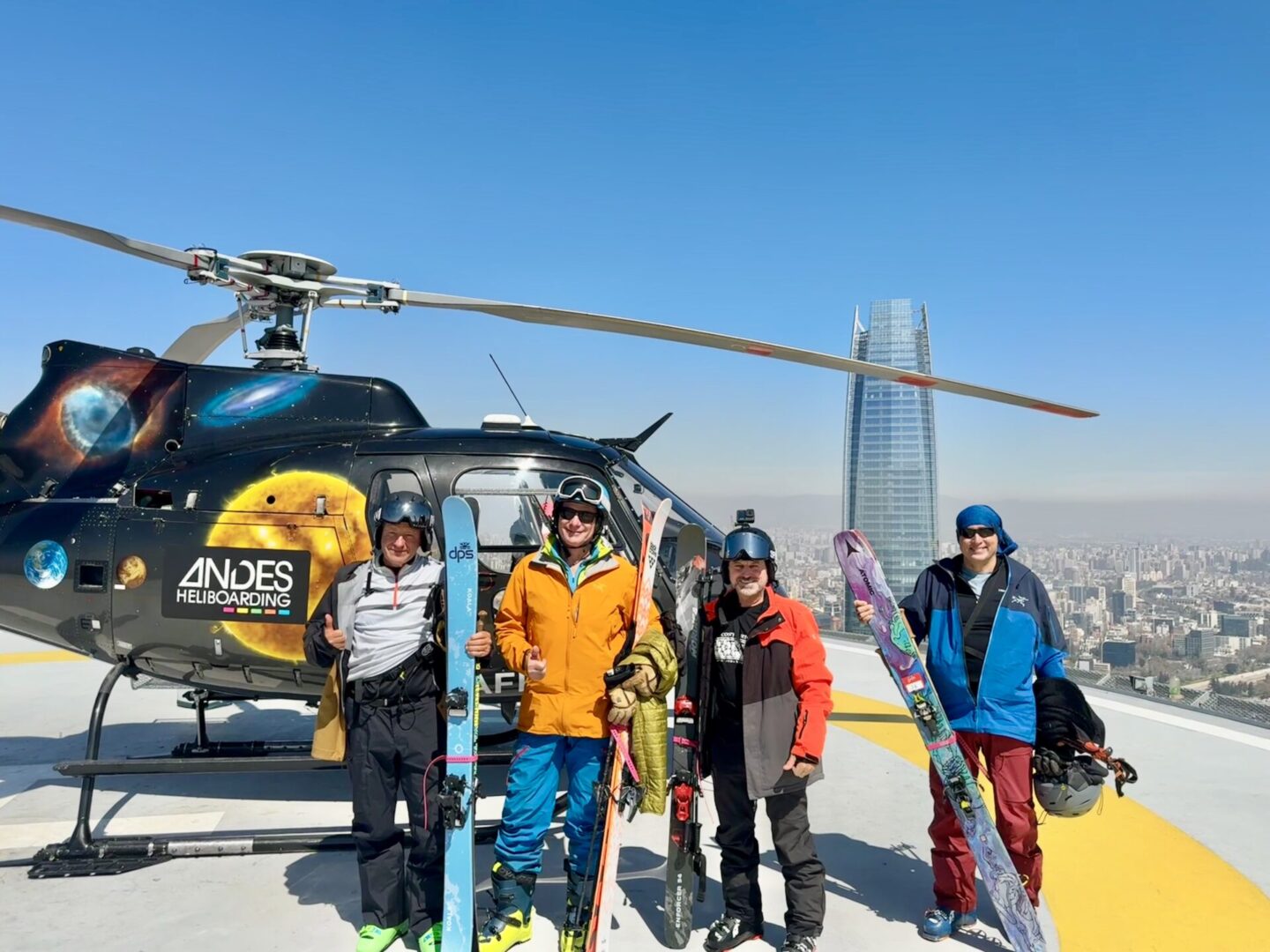 Four skiers pose with skis in front of a helicopter on a rooftop helipad.