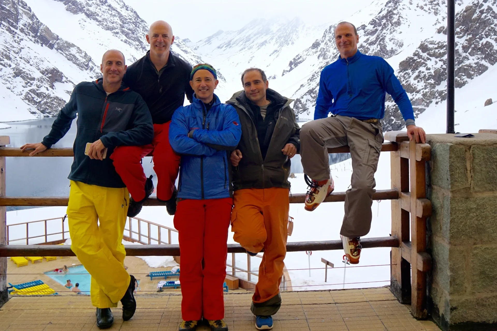 Group of skiers posing on a snowy mountain with lake and peaks behind.