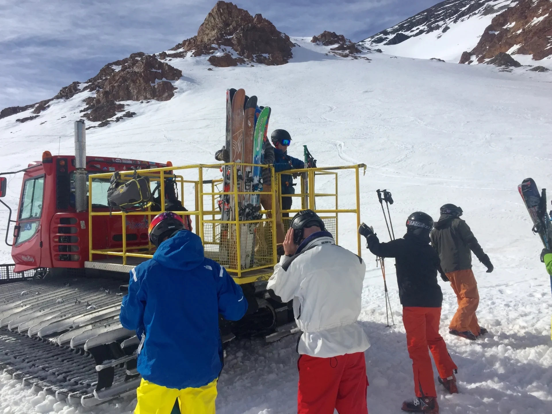 Group of skiers posing on a snowy mountain with lake and peaks behind.