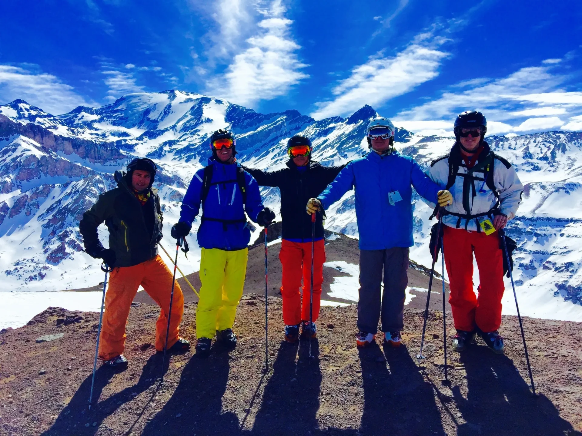Group of skiers posing on a mountain with snowy peaks behind.