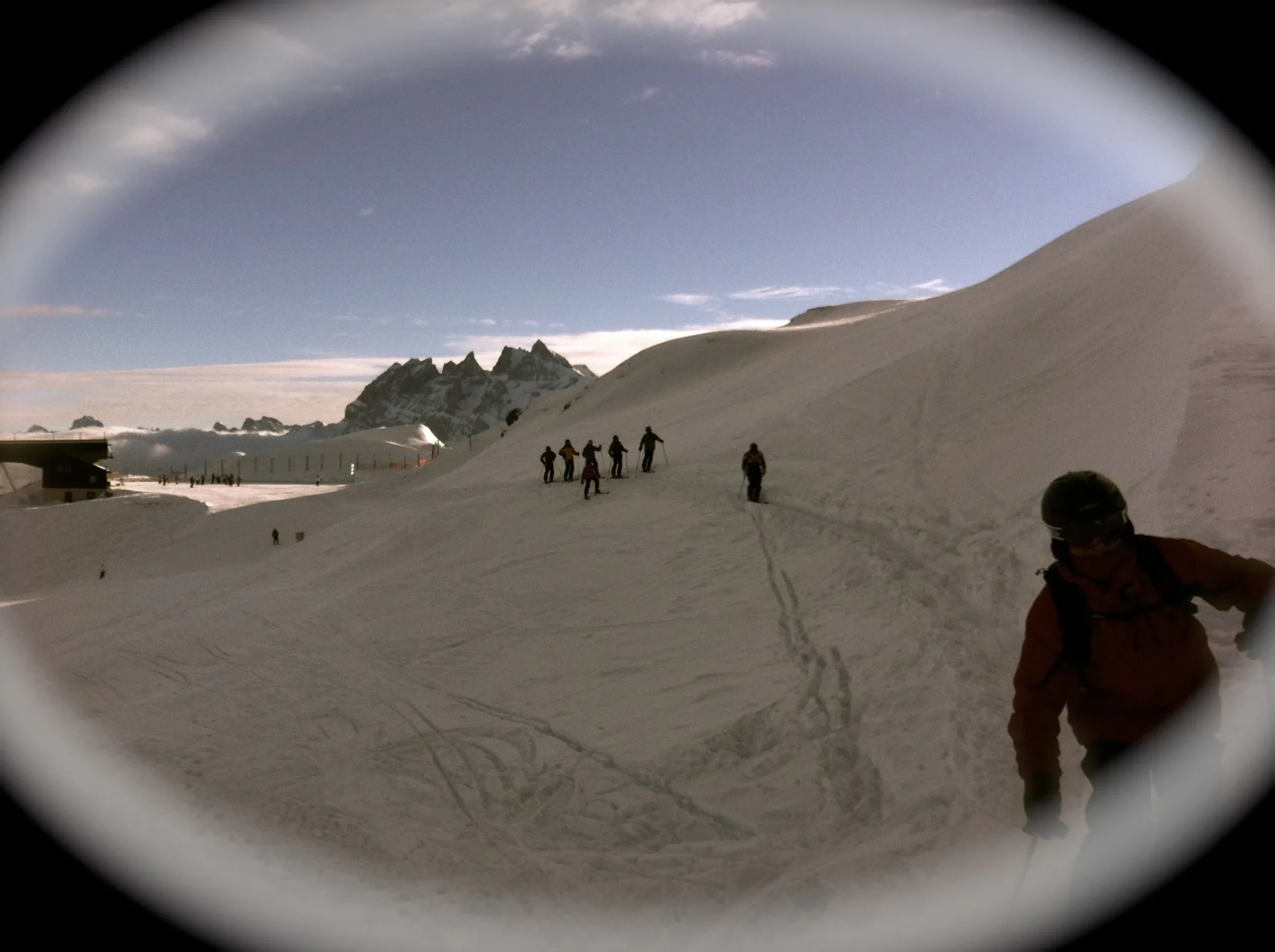 Group of skiers posing on a snowy mountain with lake and peaks behind.