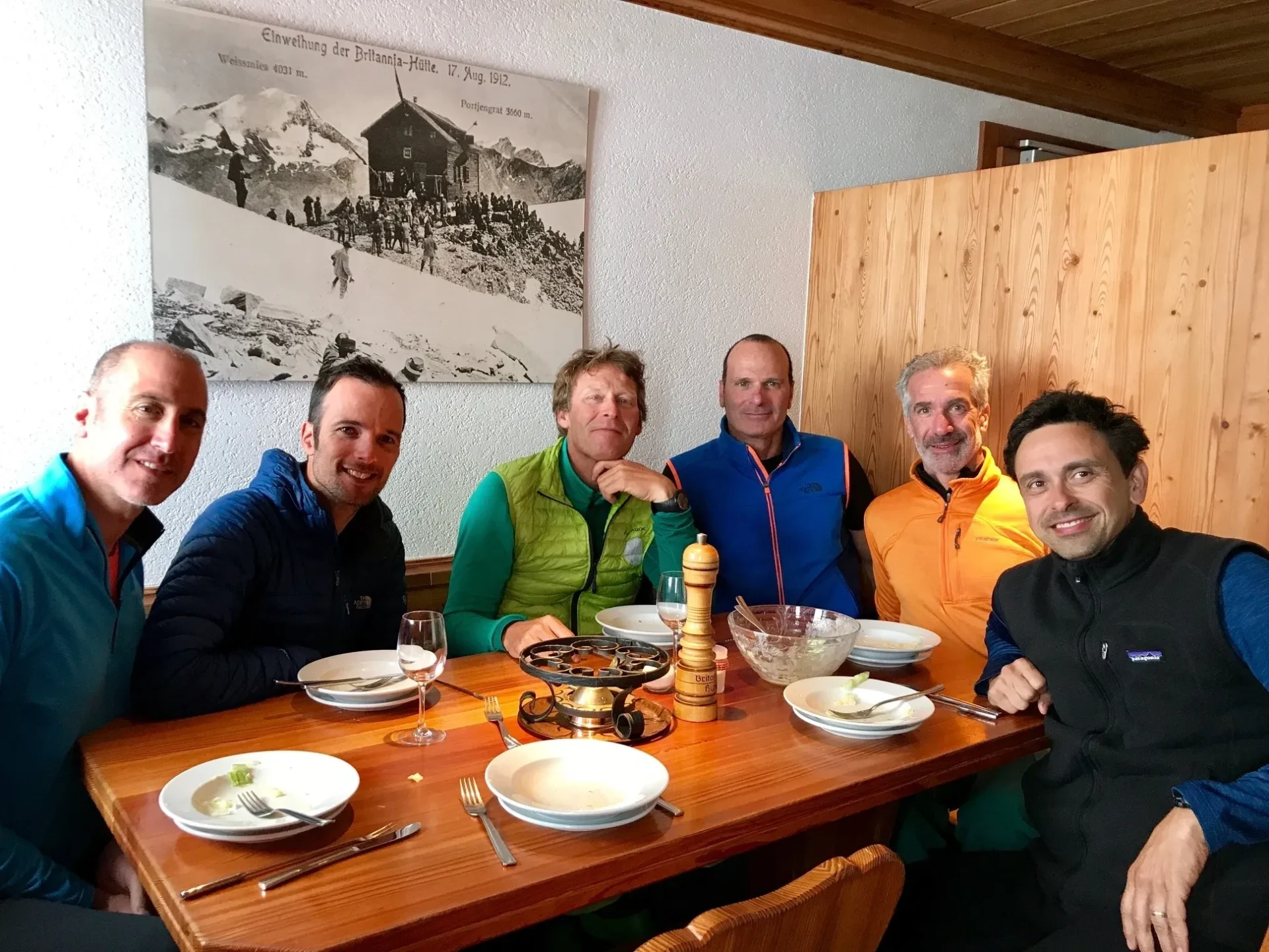 Group of skiers posing on a snowy mountain with lake and peaks behind.