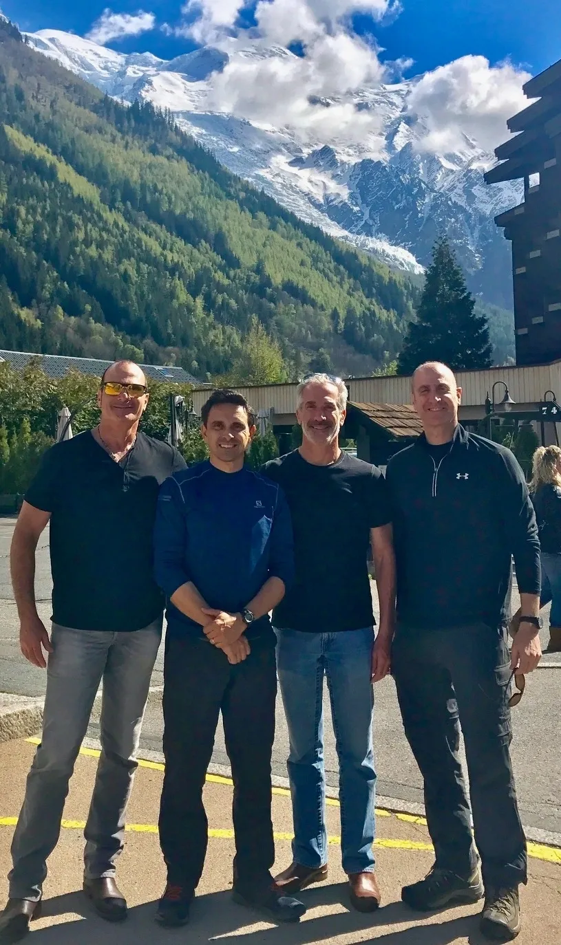 Group of skiers posing on a snowy mountain with lake and peaks behind.