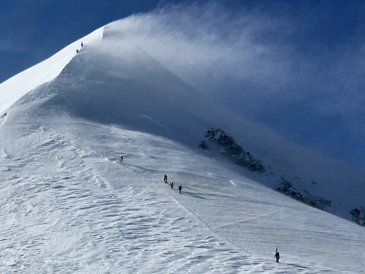 Climbers ascending a snowy mountain ridge under a clear blue sky.