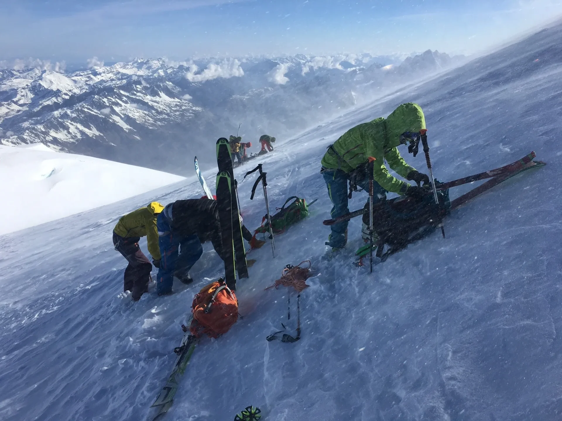 Group of skiers posing on a snowy mountain with lake and peaks behind.