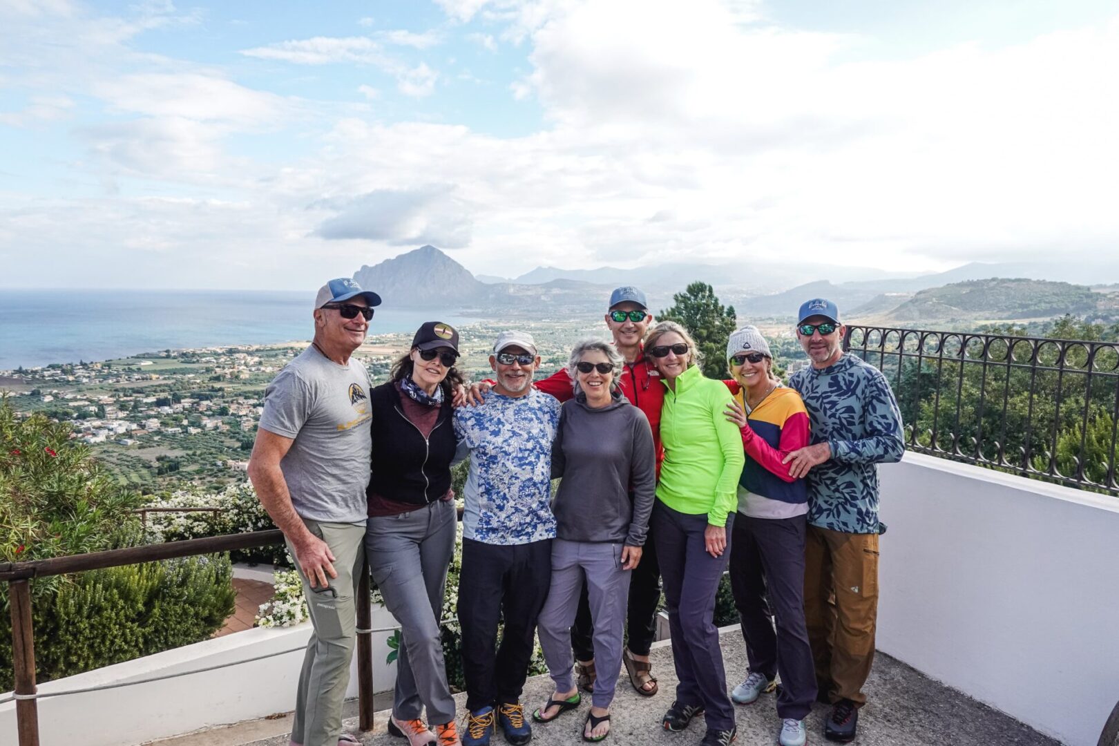 Group of skiers posing on a snowy mountain with lake and peaks behind.