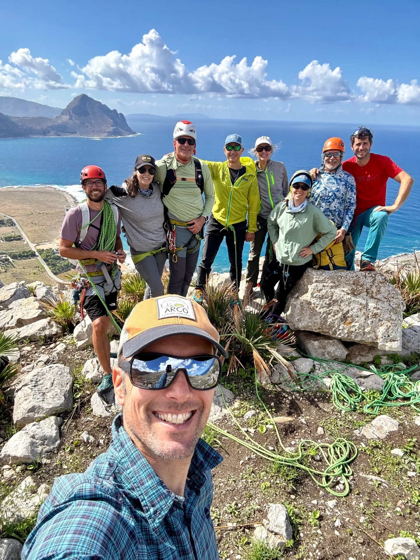 Group of skiers posing on a snowy mountain with lake and peaks behind.