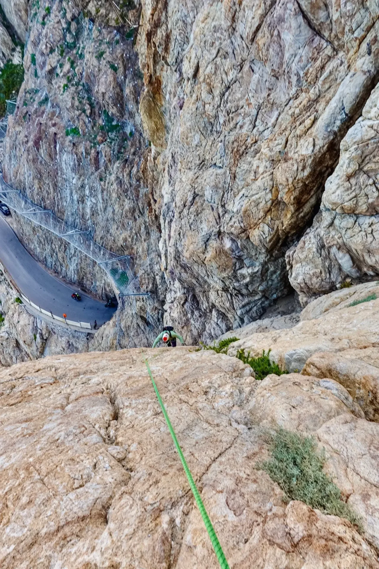 Person climbing a steep rocky cliff with safety rope and a road below.